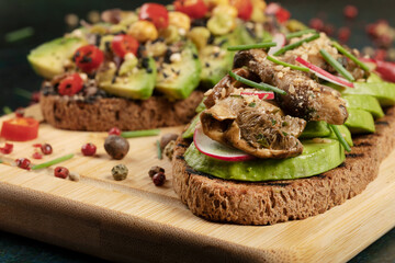 An avocado toast with mushrooms, radish, sesame seeds and chive on a wooden cutting board.