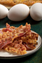 Close up of some crispy bacon slices and three fresh eggs on the background.