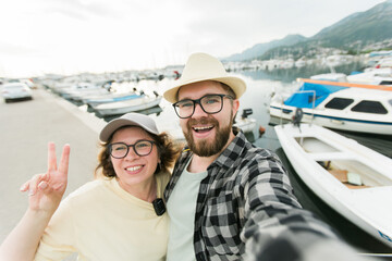 Young couple taking a self portrait laughing as they pose close together for camera on their smartphone outdoors in summer port marina with boats and yachts