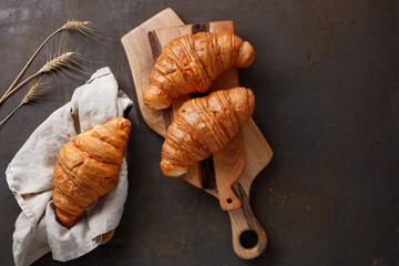 Board with tasty croissants on dark table with spikelets, close up. French pastry. Fresh croissants on dark brown background, copy space for your product. Selective focus. Top view