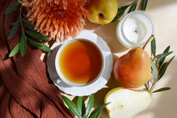 Cup of tea with croissant, pears, candle, green branches on the light brown background. Fall season tea. Sun light rays.  