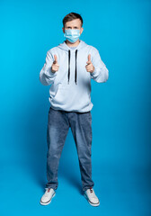 Fototapeta premium a guy in a blue sweatshirt, jeans in a medical mask. and points fingers at us. full height. blue background