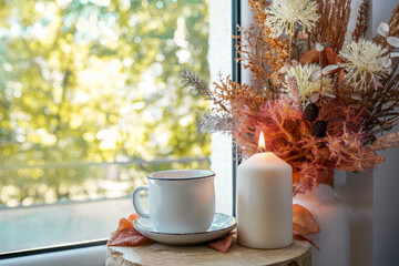 Cup of autumn tea (coffee, chocolate) and yellow dry leaves in the vase, candle near a window, copy space. Hot drink for autumn cold rainy days. Hygge concept, autumn mood