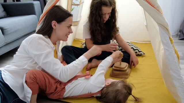 Top view of playful mother enjoying free leisure time with two little daughters. Adorable small kids girls lying in cozy tent with happy young mom playing with wooden cubes in living room at home