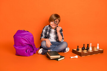 Full length photo of young boy sit floor look zoom investigate chess bag isolated over orange color background