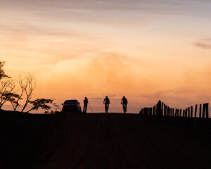 Silhouette of photographer photographing two cyclists on rural farm road, mountain biking during sunset, with red to orange sky
