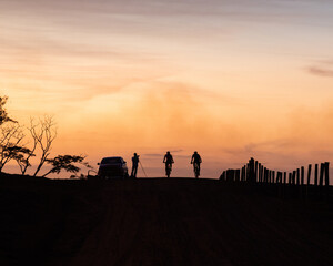 Fototapeta premium Silhouette of three cyclists on a rural road with a fence at sunset, orange-yellow sky