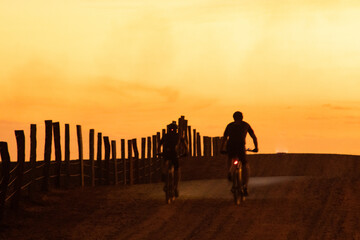 Fototapeta na wymiar Two people riding bicycles during sunset in rural area, in silhouette and sky in orange tones.