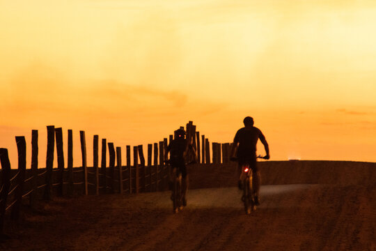Two people riding bicycles during sunset in rural area, in silhouette and sky in orange tones.