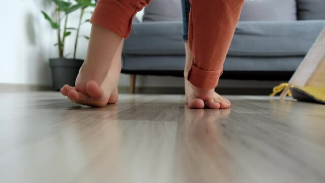 Selective focus of little daughter child staying on mother feet, learning dancing at home on warm floor. Close up of unrecognizable mom with kid girl having fun enjoy playing funny activity together