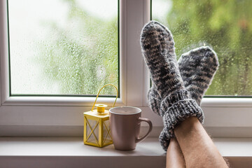 Man's feet wearing knitted wool socks near the hot cup of tea with yellow lantern on windowsill at cozy autumn home.