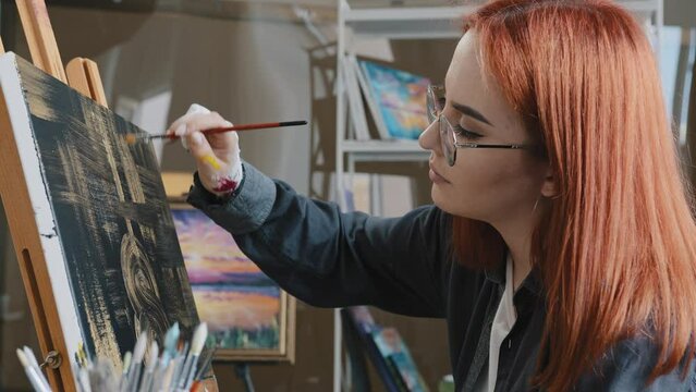 Inspired busy concentrated Caucasian red-haired woman in eyeglasses girl painter wear glasses painting drawing on canvas with yellow gold paints practicing hobby art education skill draw with brush