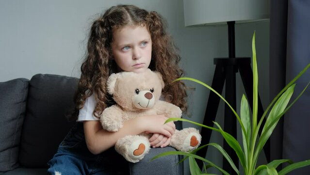 Close up of upset lonely bullied little girl child holding teddy bear toy looking away feels abandoned abused, sad unhappy small kid orphan sitting alone on grey sofa at home, charity adoption concept