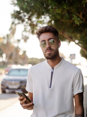 Hombre joven vistiendo gafas de sol usando su teléfono móvil en la calle durante sus vacaciones de verano