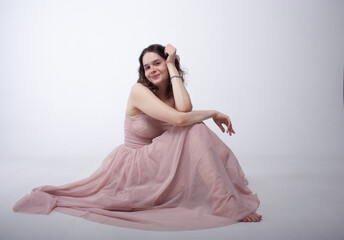 Fototapeta premium A cute girl in a pink long dress is sitting, leaning her head on her hand and smiling. The girl looks at the camera.