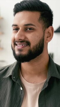 Vertical portrait of confident attractive indian or arabian man, in casual stylish clothes, stand at home in cozy living room on the background of the kitchen, looks at camera, smiling friendly