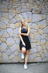 Fototapeta na wymiar Blonde young woman against stone wall background, smiling and posing on background of vineyards