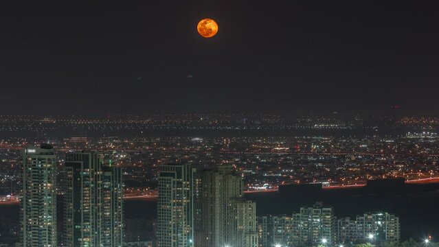 Moon rising over greens and al barsha heights district area night timelapse from Dubai marina. Towers and skyscrapers with traffic on roads from above