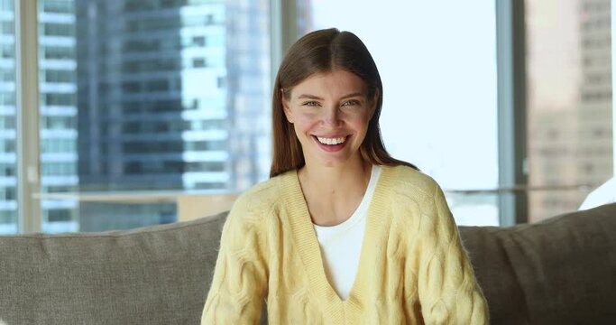 Happy beautiful woman wear yellow knitted casual sweater, rest on cozy sofa in modern skyscraper apartment, smile look at camera. Owner or renter of fashionable flat owner head shot portrait concept