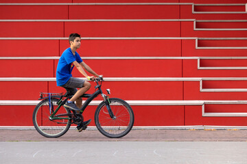 A young teen boy in a blue t-shirt rides a bicycle past a red wall