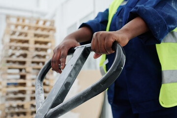 Hands of young black woman in uniform holding by handle of forklift loader with pallets while pushing it forwards during work