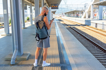 A lonely young man looks into a smartphone, dressed in a shorts T-shirt, with a backpack on his back, standing on a railway platform waiting for a suburban electric train.