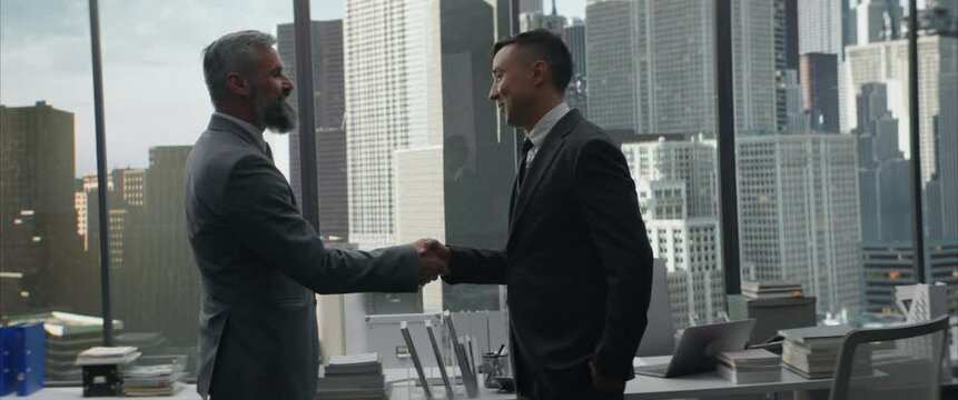 Portait of 50s Caucasian businessman shaking hands with his business partner in the office, district with skyscrapers in the background. Shot with 2x anamorphic lens