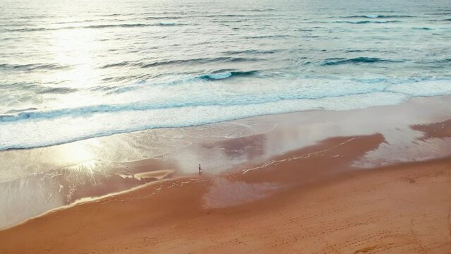 Orbit movement around tiny figure of runner on empty scenic ocean beach at low tide time with golden sunset light