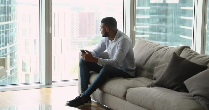 Young successful African man using smartphone rest on sofa looks at big city skyscrapers through panoramic window in living room. Lifestyle, modern technologies usage, comfort life, wellbeing concept