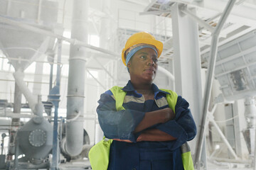 Young African American female engineer in blue uniform and yellow hardhat keeping her arms crossed on chest during work in factory