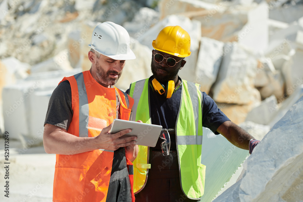 Poster two interracial male engineers in hardhats and uniform looking at tablet screen while discussing onl