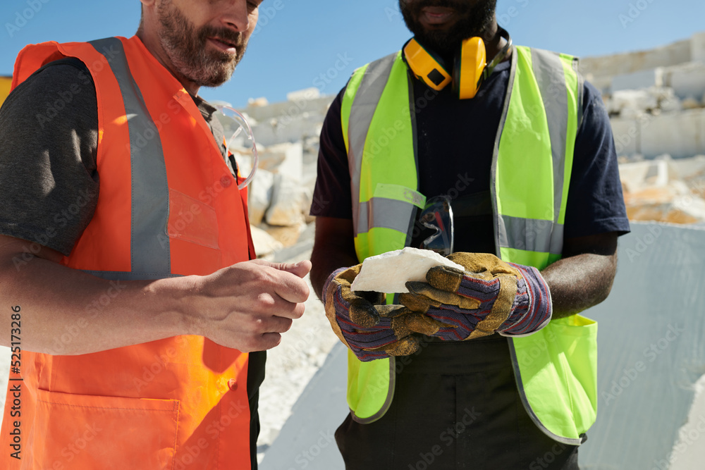 Poster mid section of two intercultural workers of marble quarry in uniform discussing sort of solid rock w