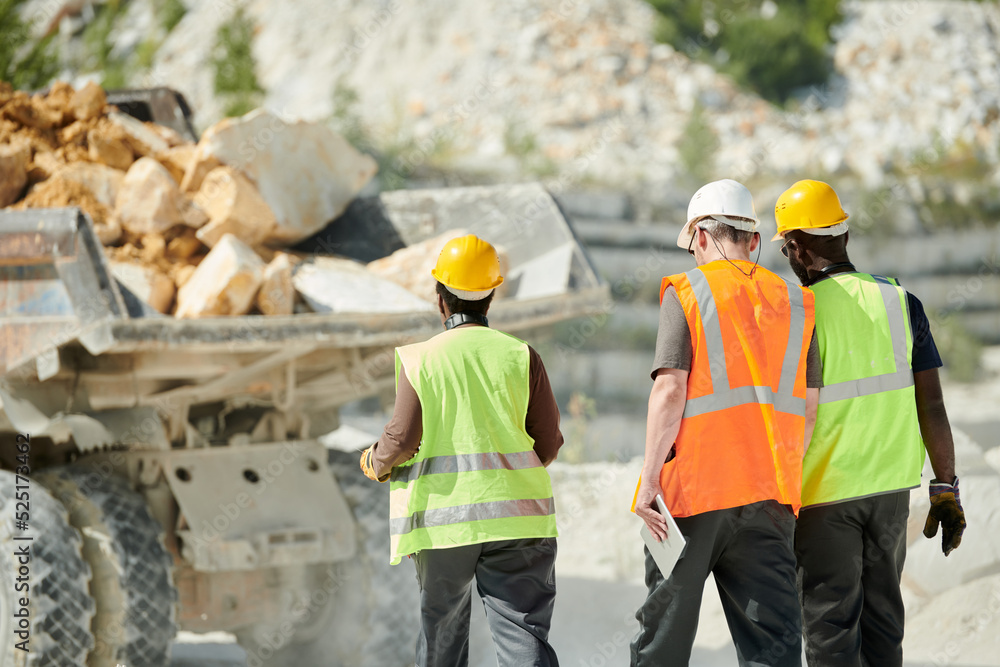 Poster rear view of intercultural engineers in uniform and hardhats walking towards dump truck with heap of