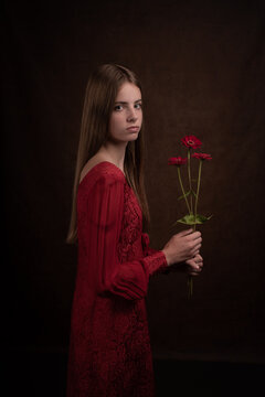 Classic dark studio portrait of young woman in red dress holding zinnia flowers
