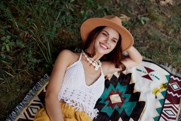 A woman in eco-friendly clothing in a hippie look is lying on a colored plaid smiling and looking at an autumn sunset in nature in the park. Lifestyle on a journey of harmony with the world