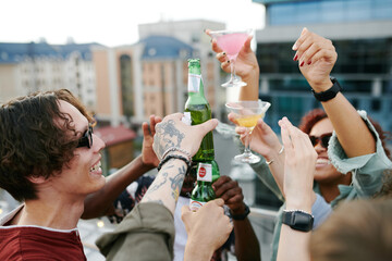 Cheerful guys and girls toasting with bottles of beers and cocktails at rooftop party while...
