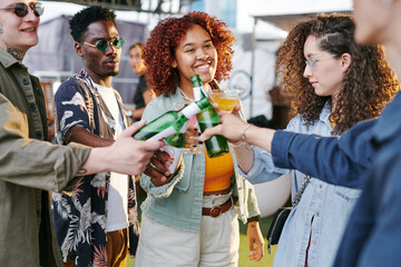 Group of happy young intercultural friends in casualwear toasting with beer and cocktails and enjoying rooftop party on weekend