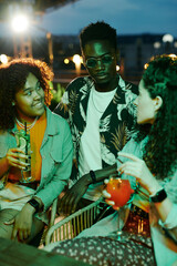 Two intercultural girls with cocktails looking at African American guy in stylish casualwear and sunglasses sitting between them