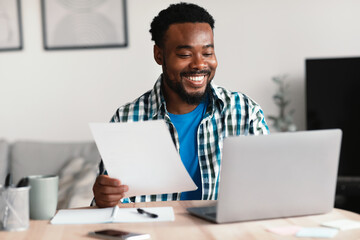 Cheerful African American Freelancer Man Using Laptop Holding Papers Indoor