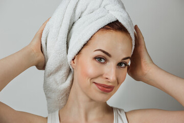 Portrait of smiling caucasian woman with healthy skin in towel posing over grey background. Happy young lady touching her perfect face with hands.