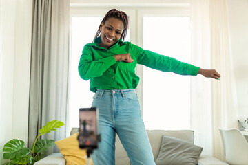 Playful african american lady dancing at camera, filming video on cellphone on tripod at home, creating trendy content