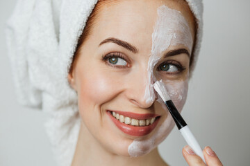 Attractive young lady smiling and looking at camera with white clay mask on half of face. Caucasian woman using brush for applying cosmetic against grey background.