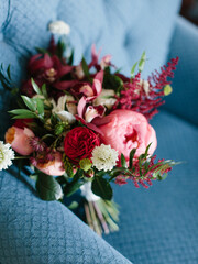 A bright bridal bouquet of pink and red peonies, white carnations, cream roses, leaves, and petals stands on a blue armchair.