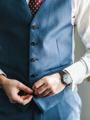 A man in a white shirt, black wristwatch, and red tie fastens a button on a dark blue vest.