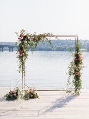A golden square wedding arch decorated with white, pink and red peonies stands on a wooden pier. Below are bouquets of flowers and decorative golden cubes. In the background there is a river.