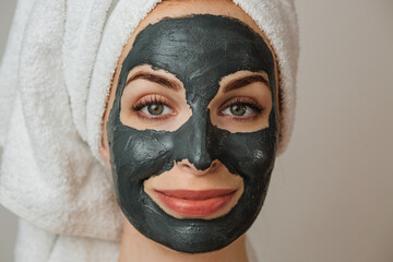 Portrait of young caucasian woman in towel with black clay cosmetic mask on her face against grey background. Regular beauty treatment for skin care.