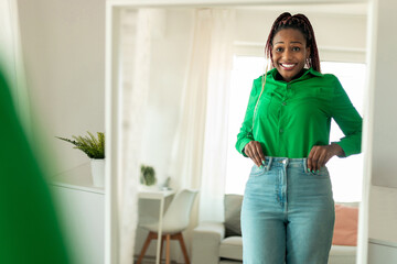 Happy african american lady after great weight loss, wearing jeans and looking at her reflection in...