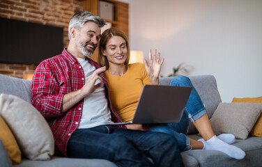 Smiling mature caucasian couple waving hand in laptop, have meeting together, sitting on sofa