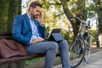 Obraz premium Tired and with headache young businessman sitting on city park bench and doing video call with associates. Bicycle in background.
