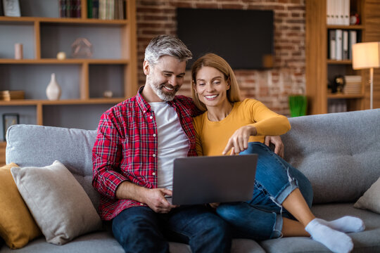 Cheerful mature caucasian wife show to man to laptop, watching video together, sit on sofa
